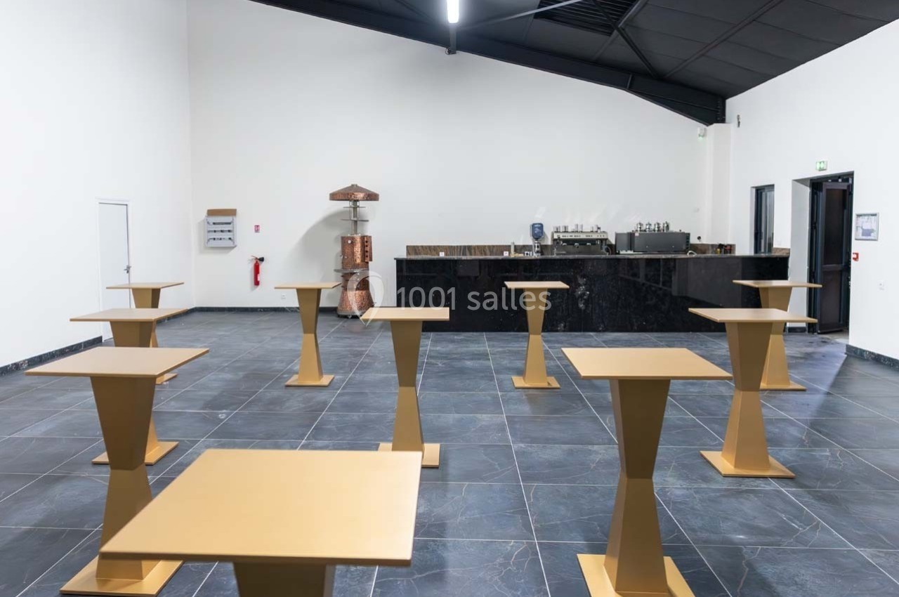 Salle vide avec des tables hautes dorées espacées, un comptoir noir en arrière-plan et un plafond incliné.