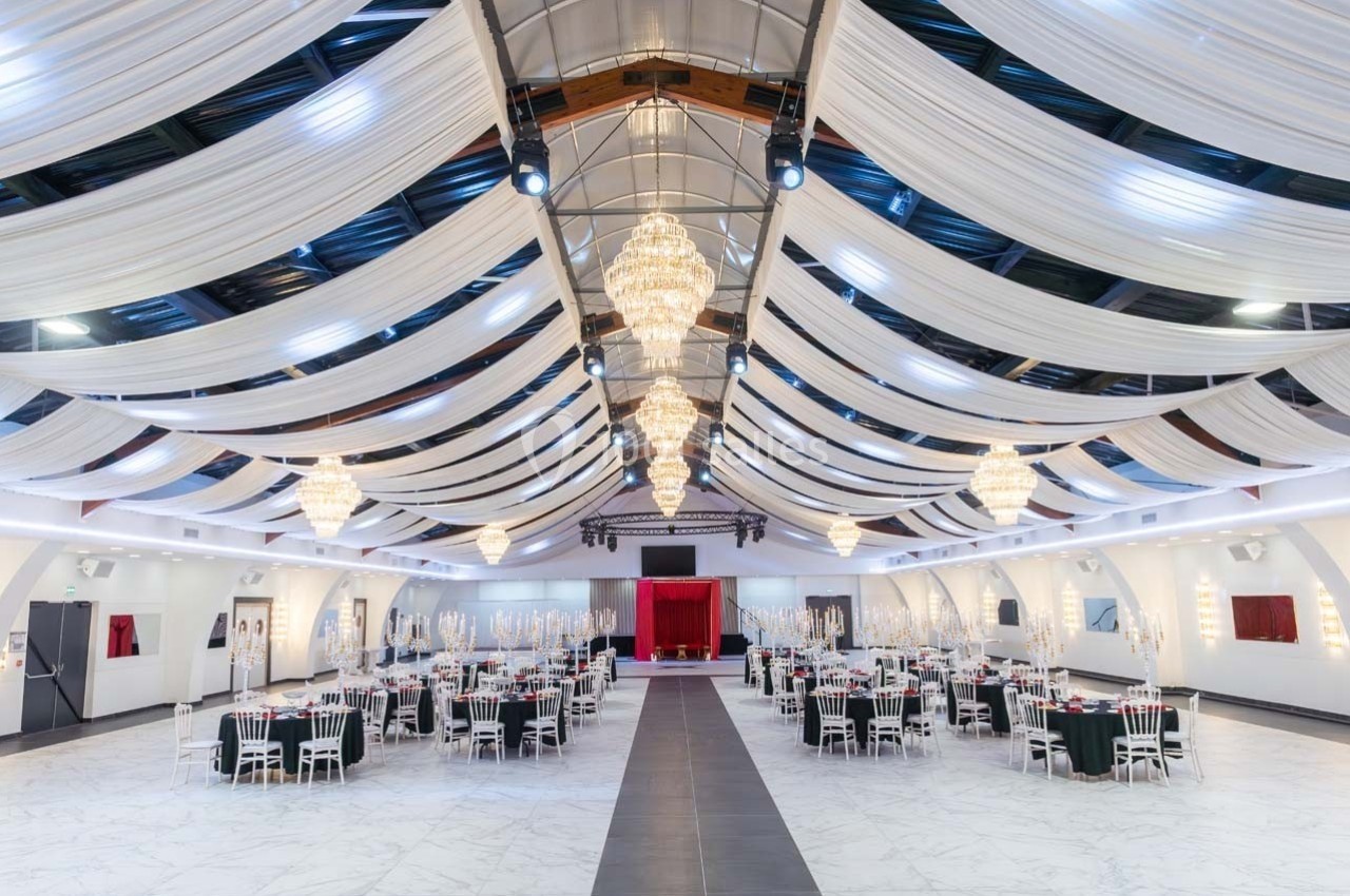 Salle de réception spacieuse avec tables dressées, lustres suspendus et plafond décoré de drapés blancs.