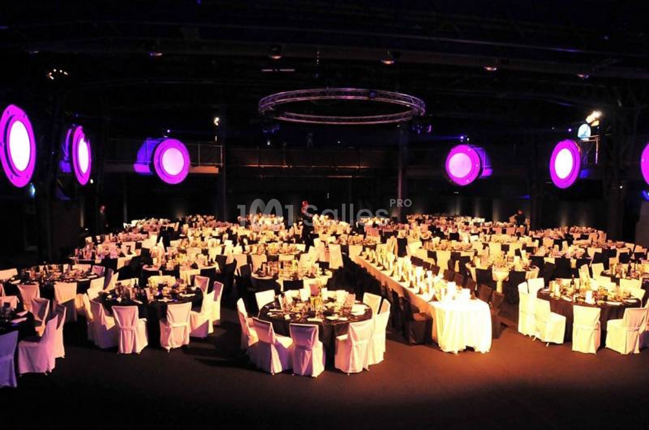 Salle de réception avec de nombreuses tables rondes dressées, éclairée par des lumières violettes suspendues.
