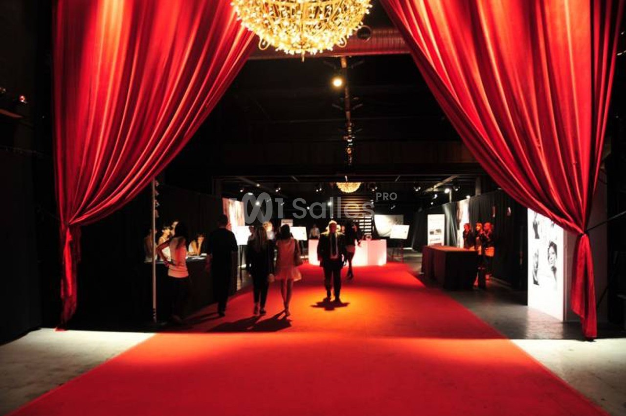 Salle sombre avec tapis rouge, rideaux rouges ouverts et lustre, accueillant des personnes lors d'un événement.