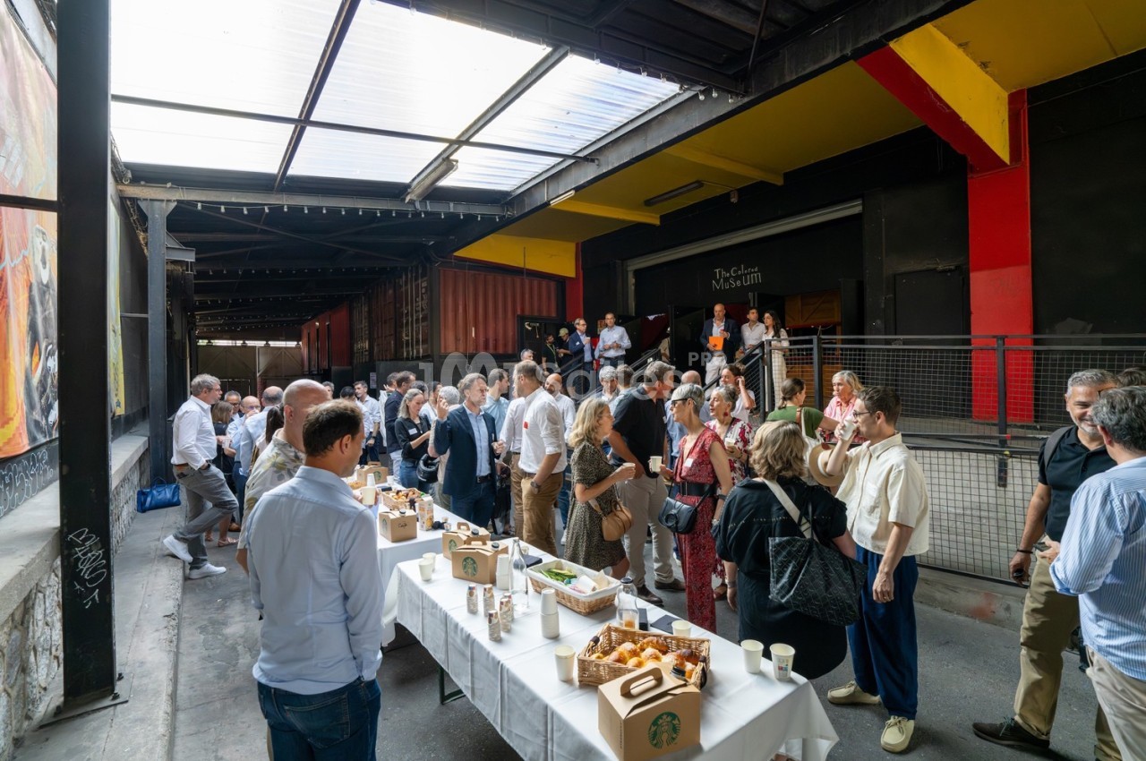 Groupe de personnes rassemblées autour de tables avec nourriture et boissons dans un espace extérieur couvert.