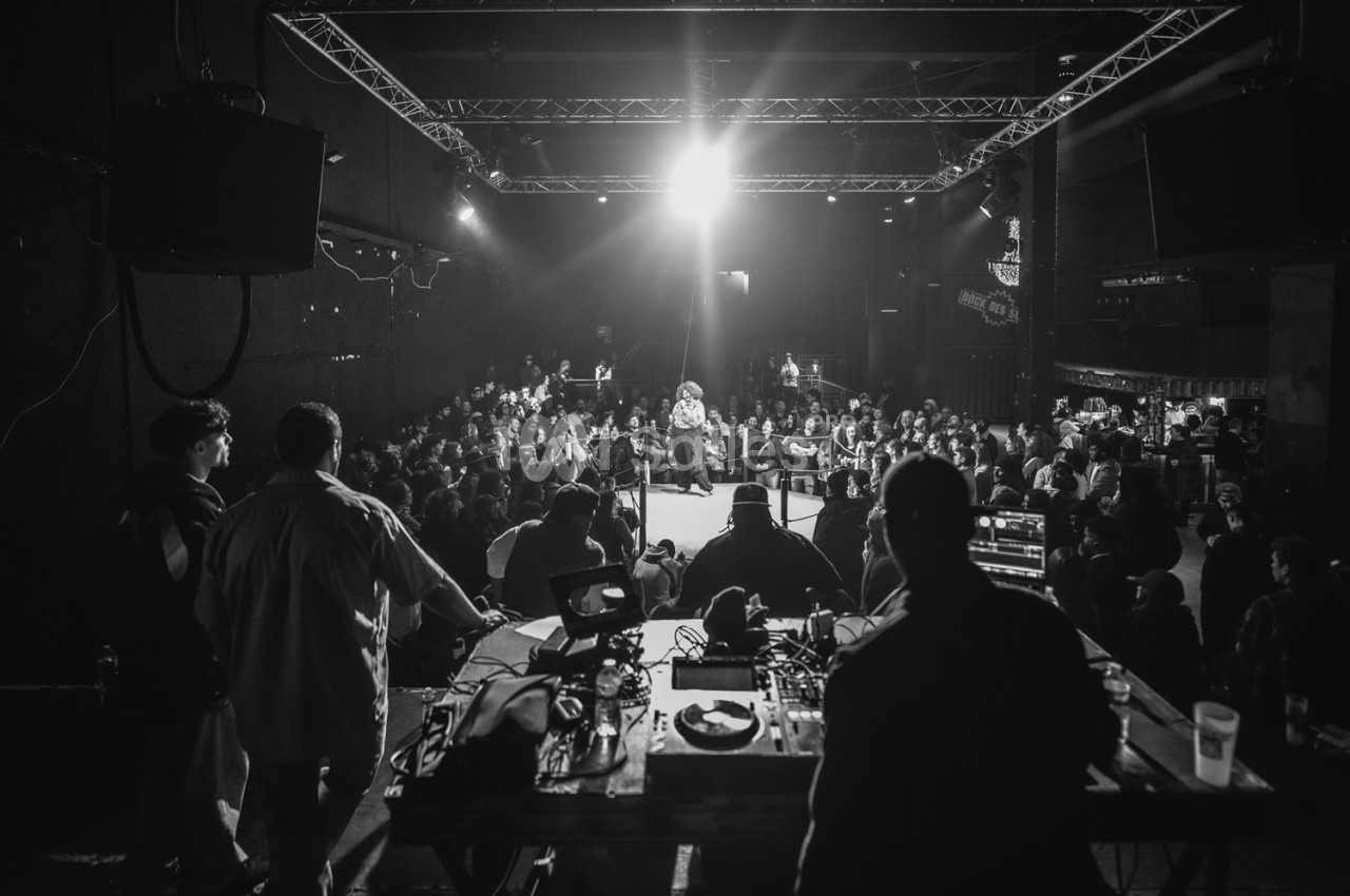 Vue en noir et blanc d'une salle de concert avec un artiste sur scène face à un public attentif.