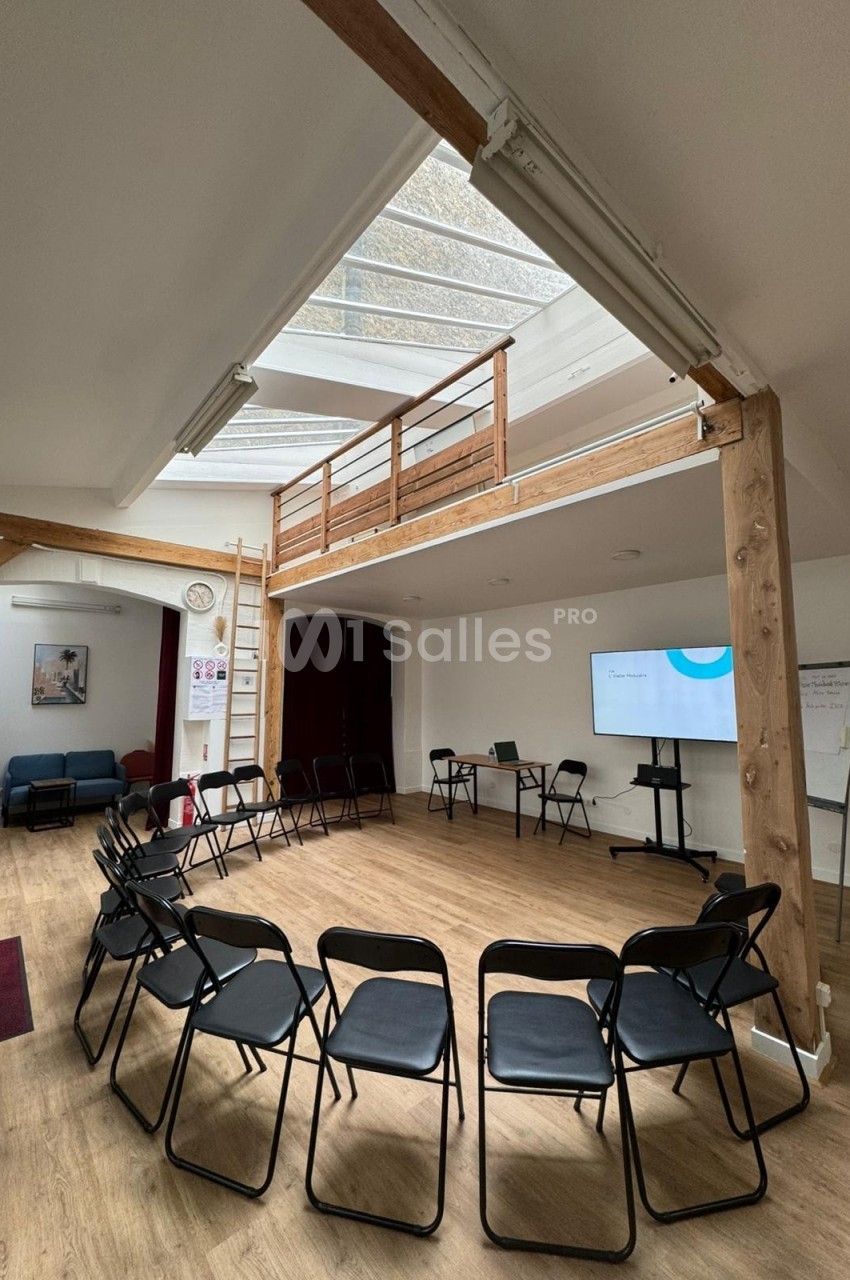 Salle lumineuse avec chaises disposées en cercle, écran de présentation, tableau blanc et mezzanine en bois.