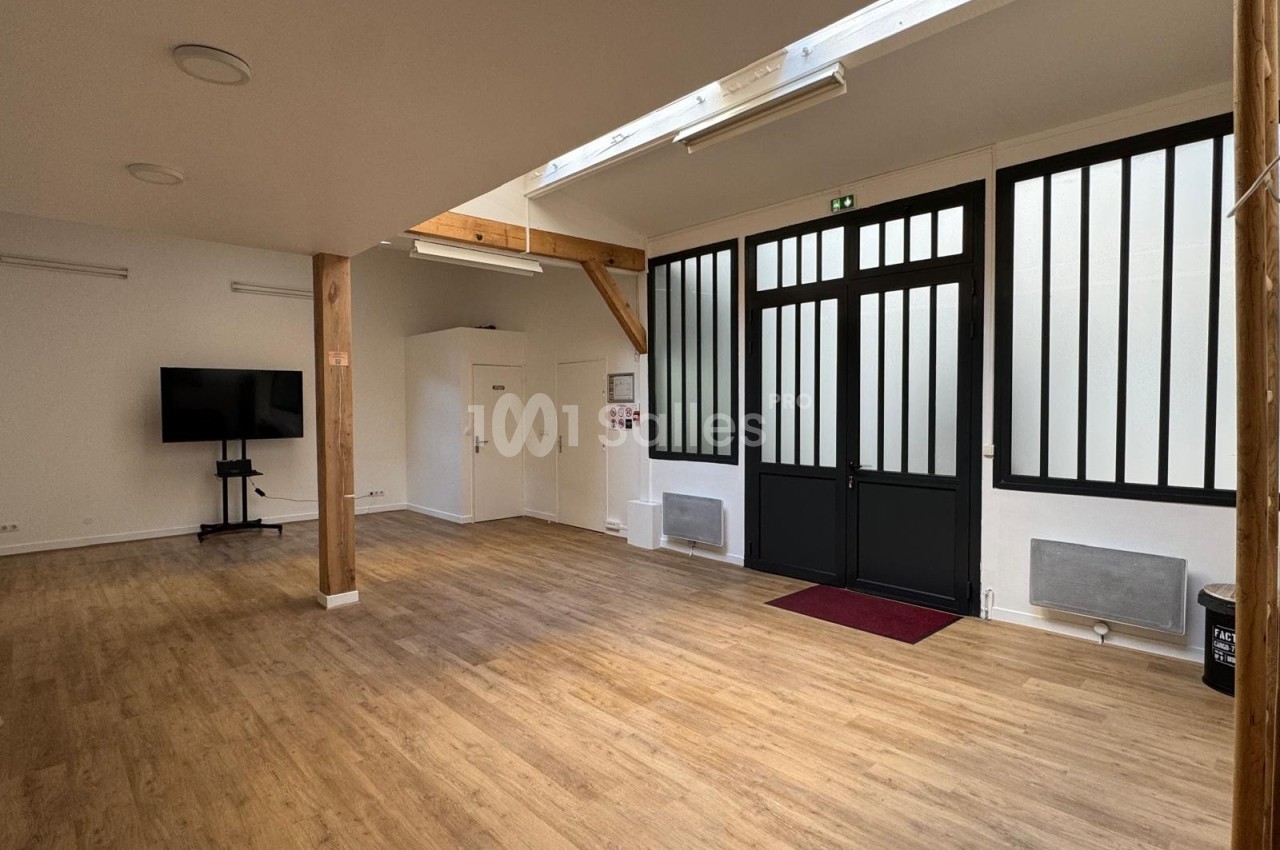Salle lumineuse avec parquet en bois, grandes portes vitrées noires, écran sur pied et poutres apparentes.