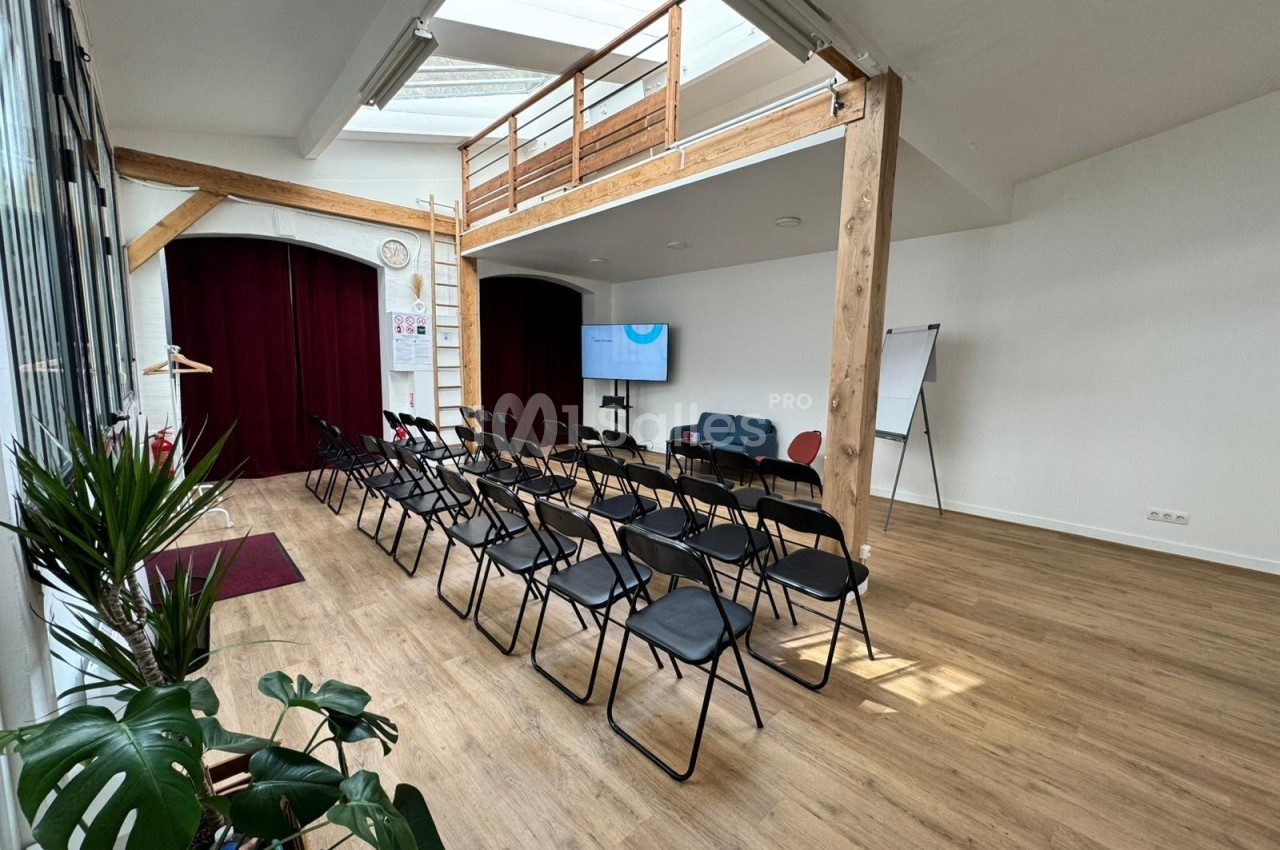 Salle lumineuse avec parquet, chaises alignées face à un écran, mezzanine en bois et grandes baies vitrées.