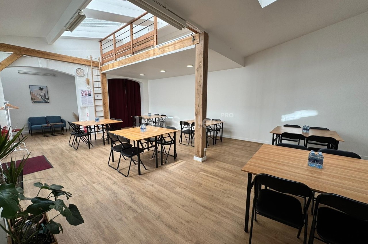 Salle lumineuse avec parquet, tables en bois, chaises noires, mezzanine en bois et plantes en décoration.