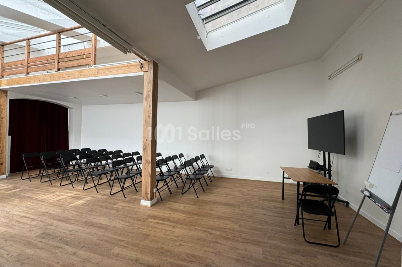 Salle lumineuse avec parquet, chaises alignées, écran, tableau blanc et mezzanine visible en arrière-plan.
