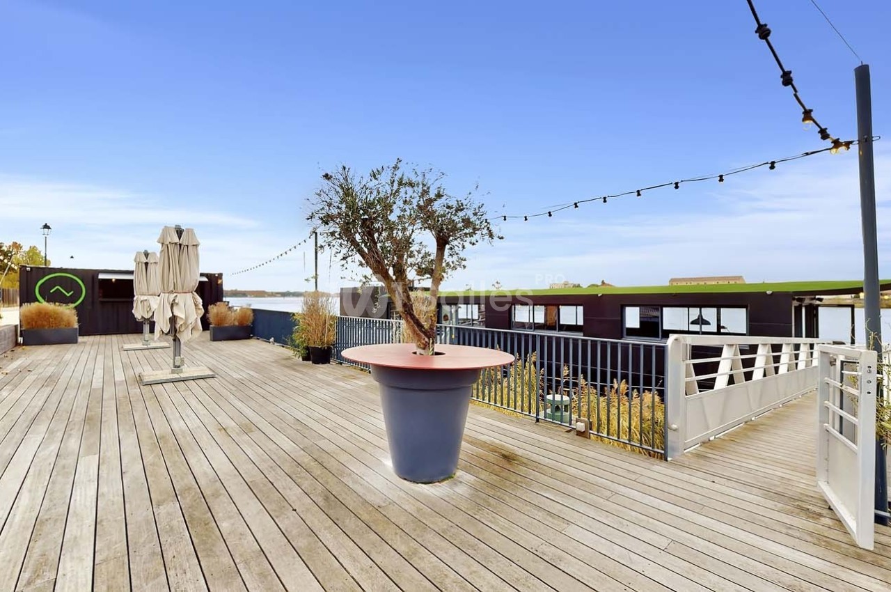 Terrasse en bois avec un grand pot d'arbre, des parasols fermés, des guirlandes lumineuses et vue sur un plan d'eau.