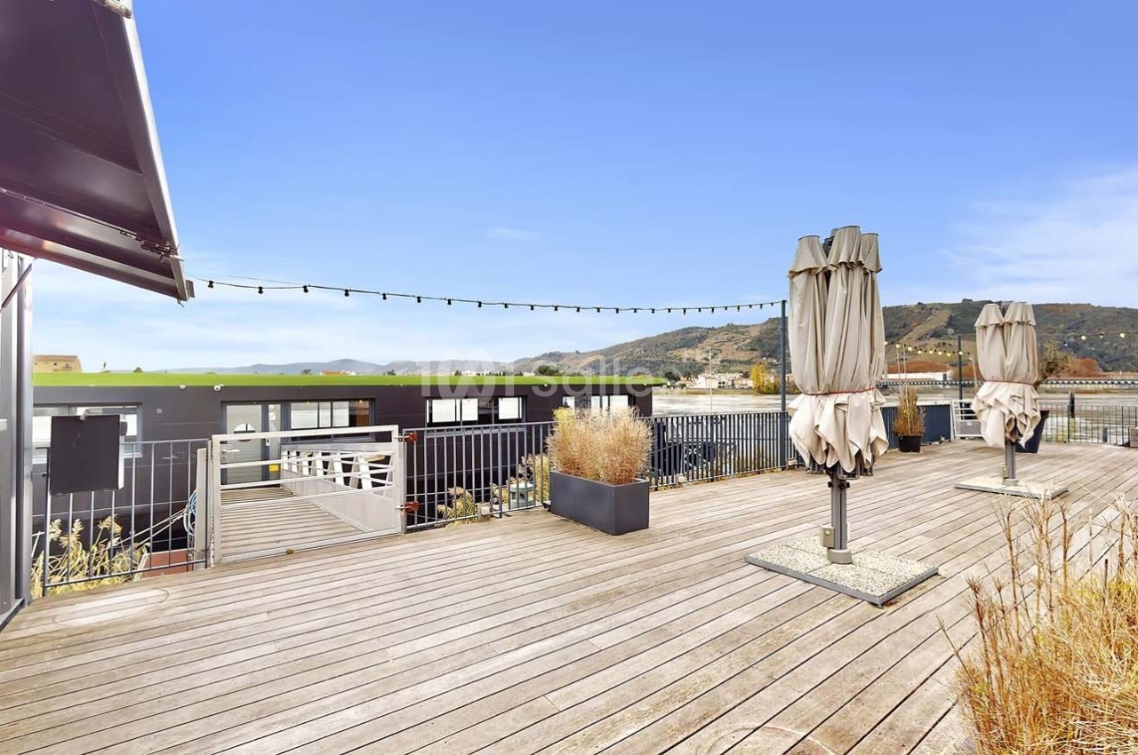 Terrasse en bois avec parasols fermés, plantes en pots et vue sur des collines sous un ciel dégagé.