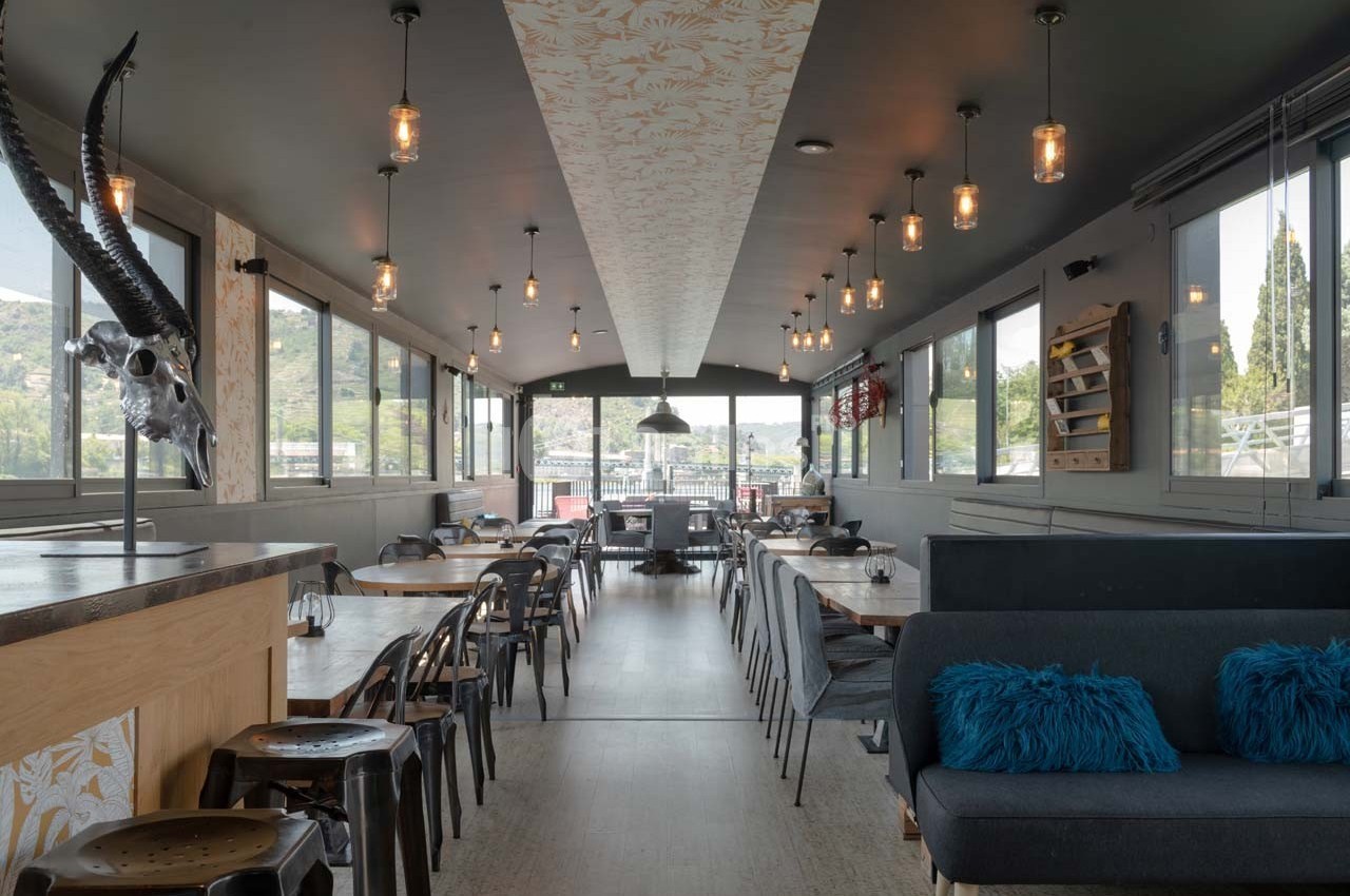 Salle de restaurant lumineuse avec grandes baies vitrées, mobilier en bois et métal, et décoration moderne.