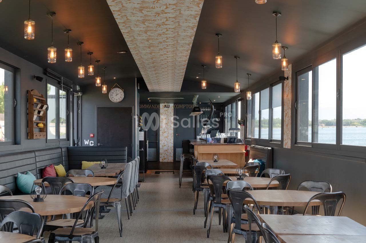 Salle de restaurant lumineuse avec tables en bois, chaises métalliques, banquettes et grandes fenêtres donnant sur l…
