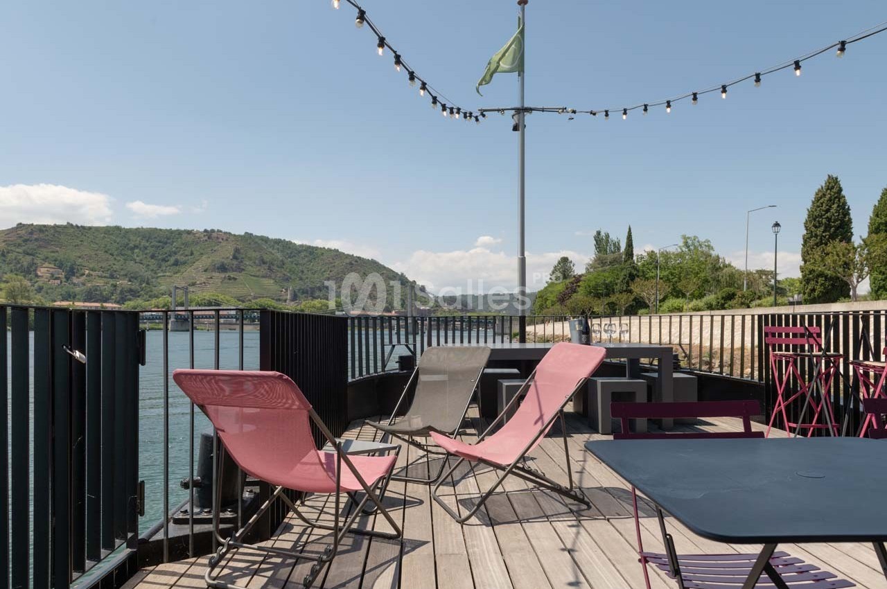 Terrasse en bois avec chaises longues et tables, vue sur une rivière et des collines sous un ciel dégagé.