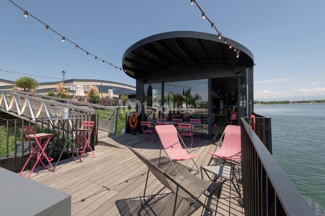 Terrasse en bois avec chaises roses, tables et guirlandes lumineuses, donnant sur un lac par une journée ensoleillée.