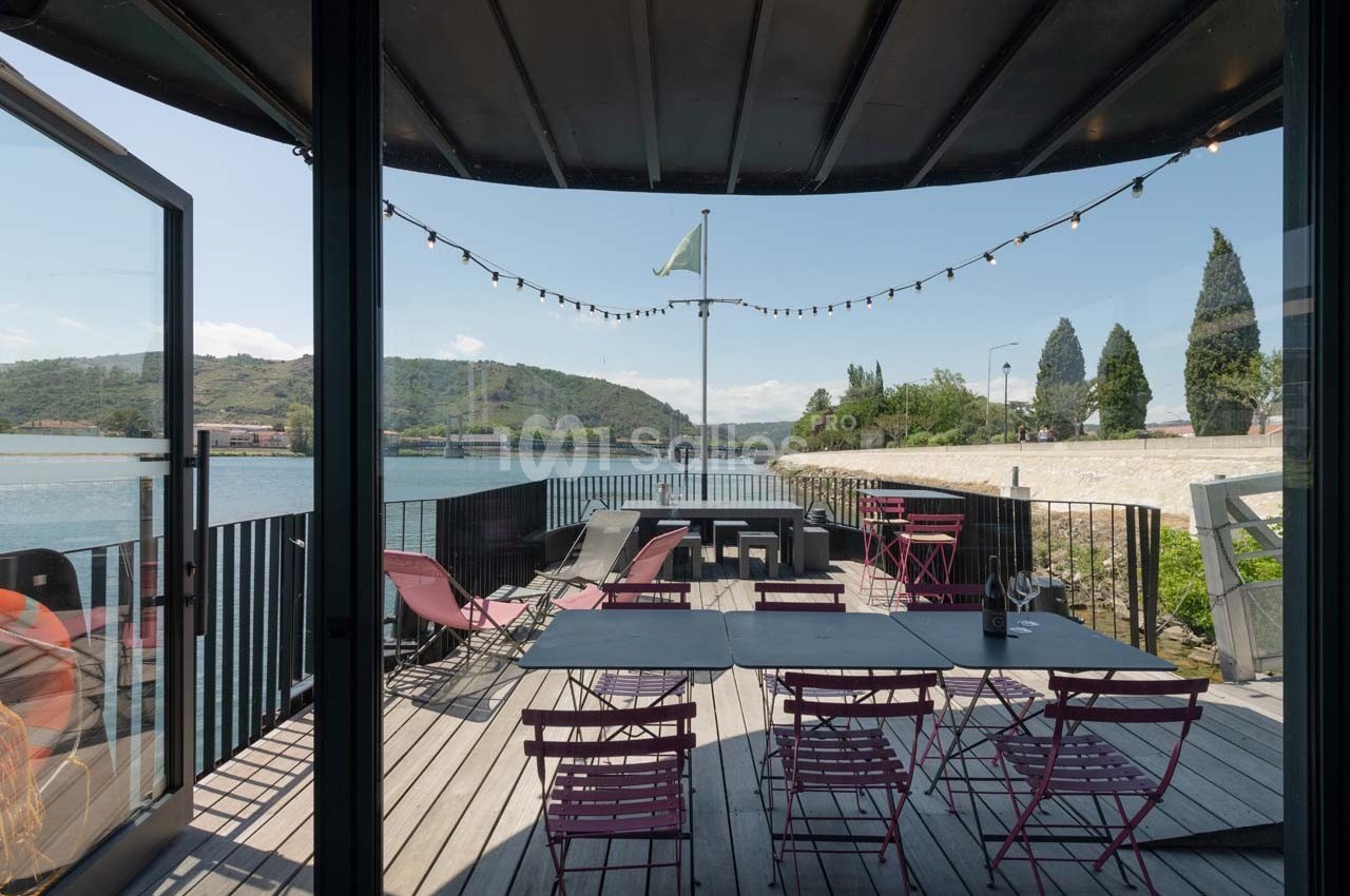 Terrasse en bois avec tables et chaises colorées, vue sur une rivière et collines sous un ciel dégagé.