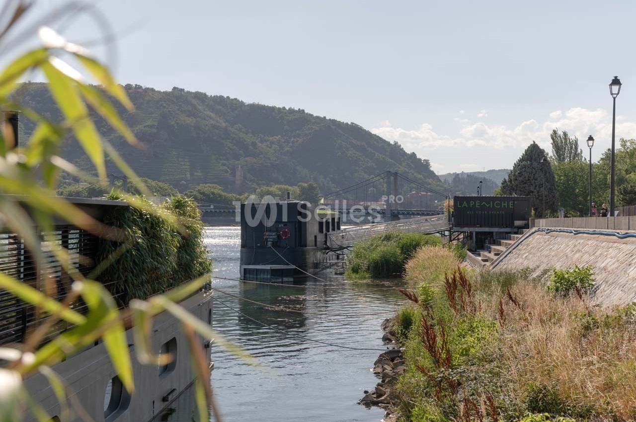 Vue d'une rivière bordée de végétation, avec une péniche amarrée et un pont visible à l'arrière-plan.
