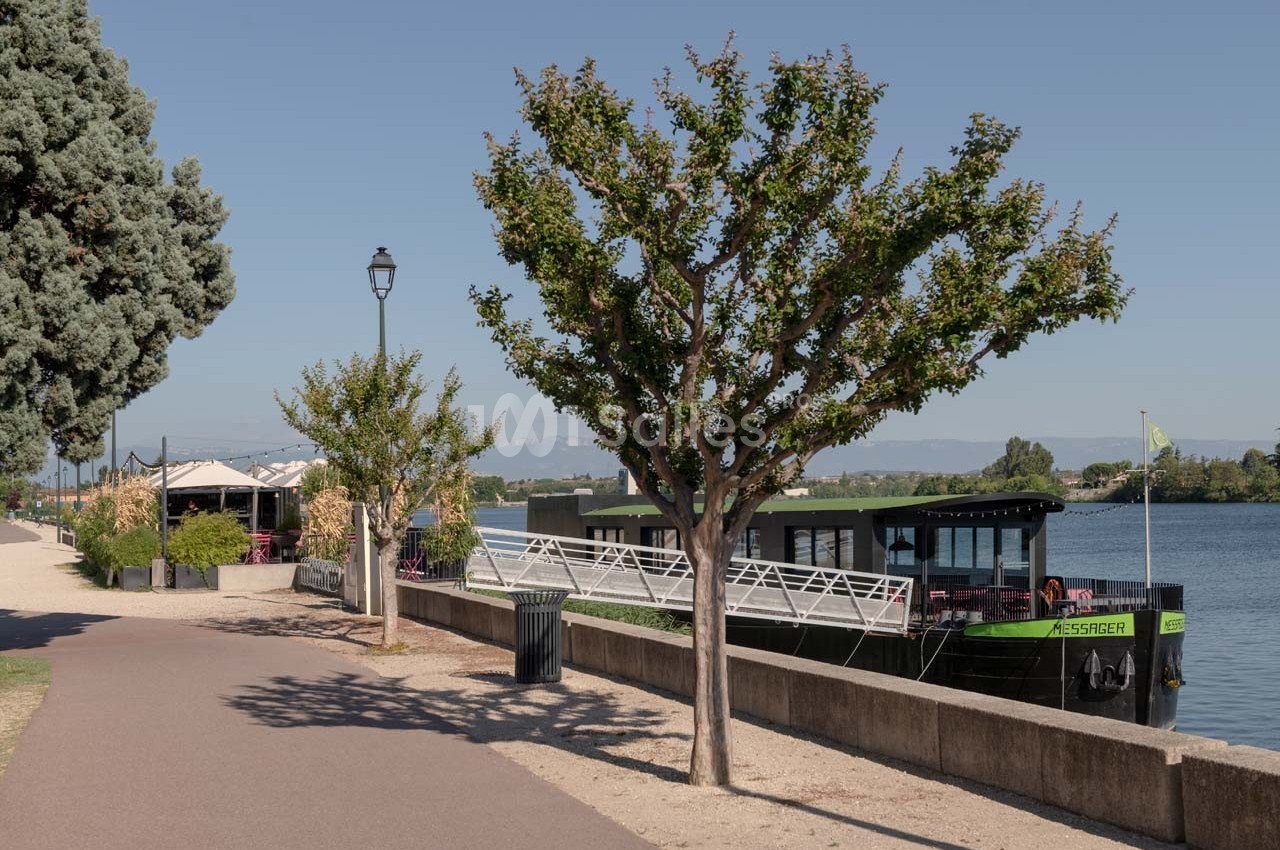 Promenade bordée d'arbres longeant un fleuve avec une péniche amarrée et une terrasse de café à proximité.