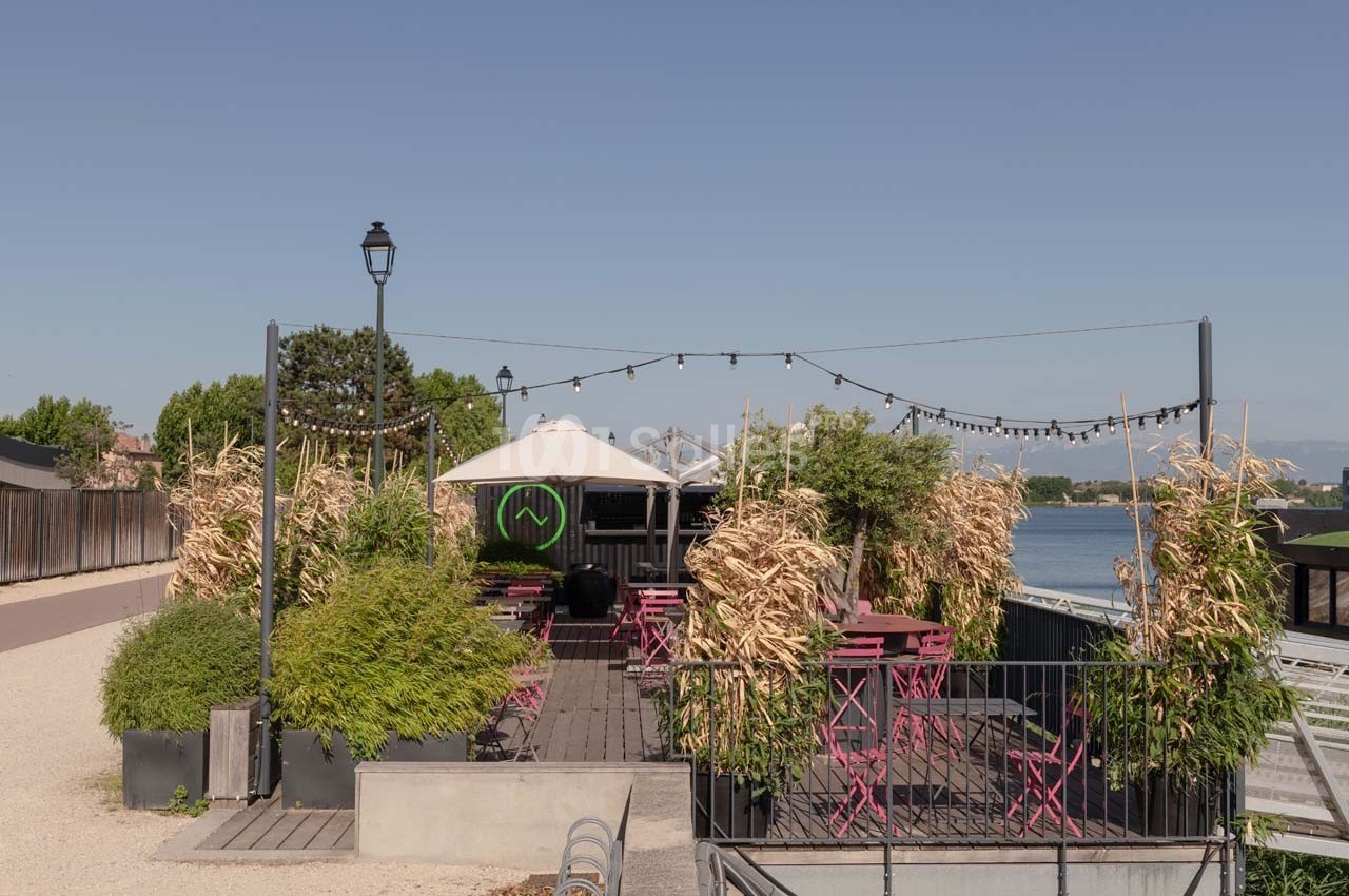 Terrasse extérieure avec tables et chaises roses, entourée de végétation, située au bord d'un plan d'eau.