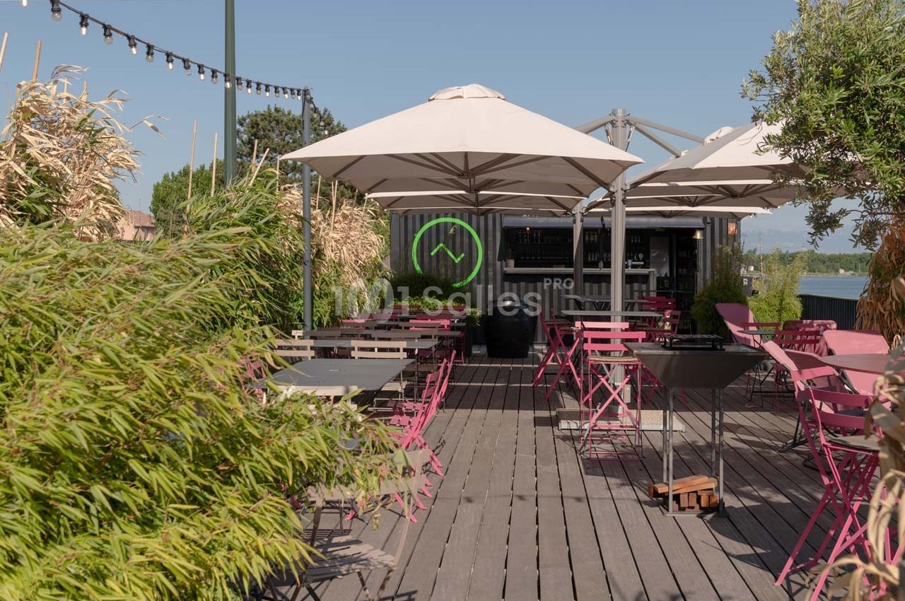 Terrasse en bois avec tables et chaises roses, parasols blancs et un bar en arrière-plan, entourée de végétation.