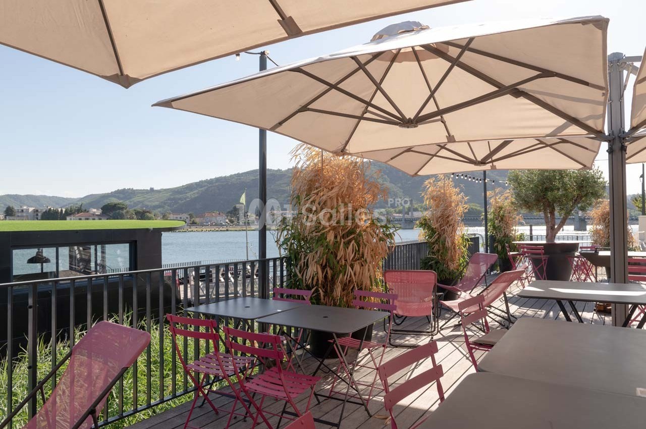 Terrasse ensoleillée avec tables et chaises colorées, vue sur un paysage de colline et un plan d'eau en arrière-plan.