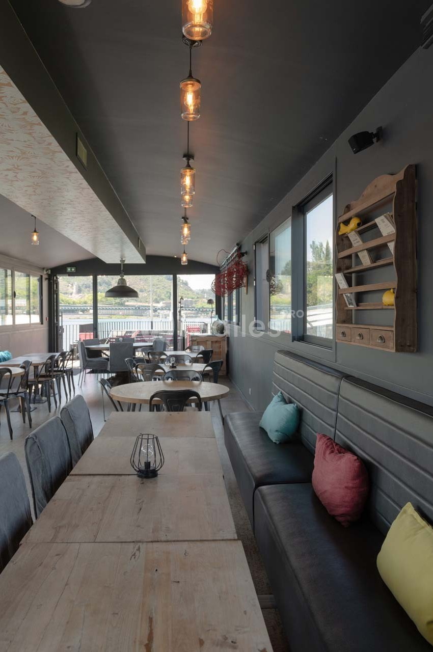 Salle de restaurant lumineuse avec tables en bois, banquettes grises, coussins colorés et suspensions industrielles.