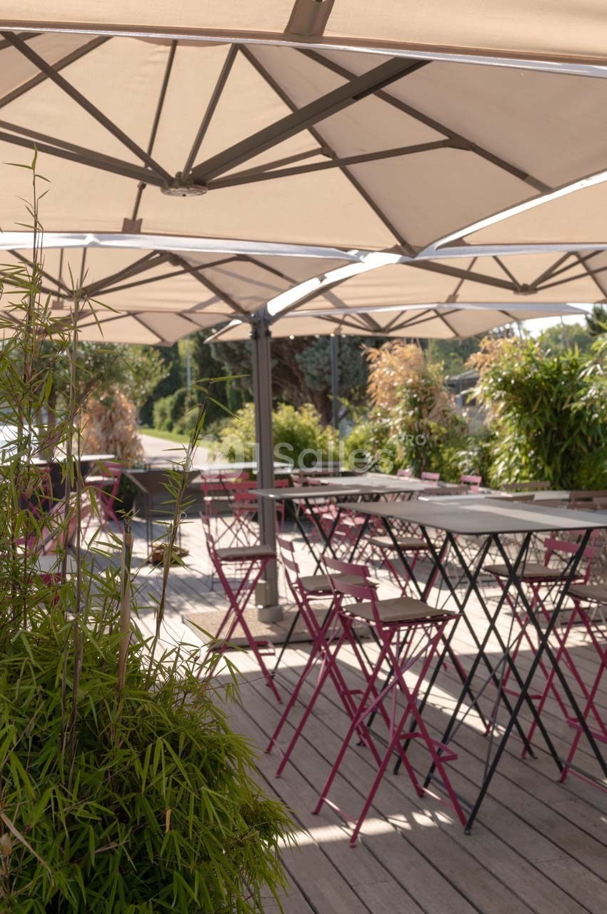 Terrasse en bois avec tables et chaises pliantes roses sous de grands parasols, entourée de plantes vertes.