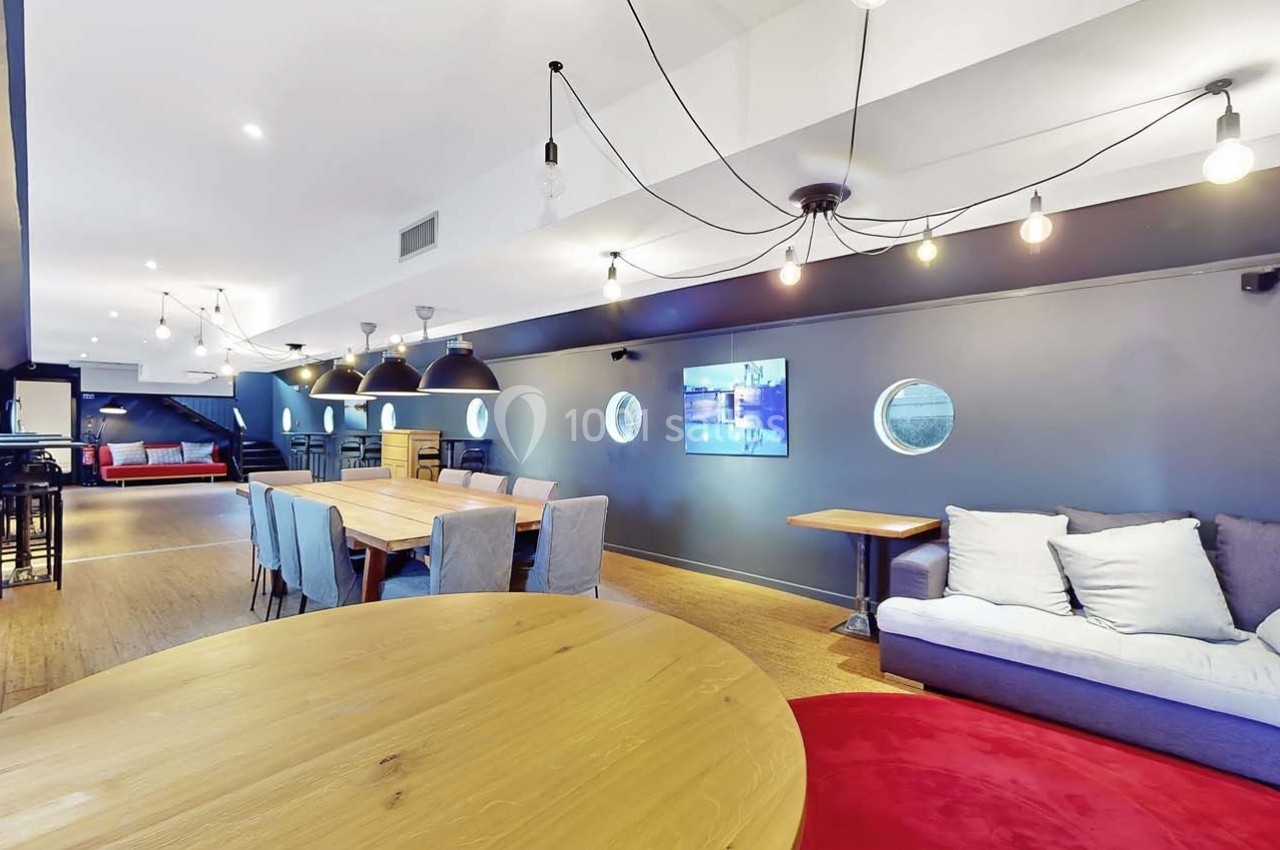 Salle moderne avec tables en bois, canapés, éclairage suspendu et murs gris ornés de hublots.