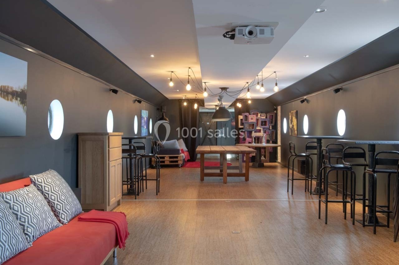 Salle intérieure moderne avec parquet, mobilier varié, éclairage suspendu et hublots sur les murs latéraux.