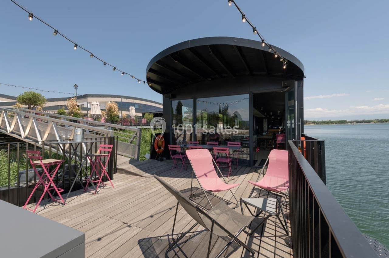 Terrasse en bois avec tables et chaises roses, située au bord d'un plan d'eau, sous des guirlandes lumineuses.