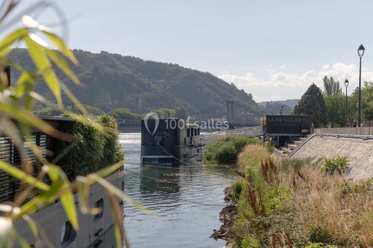 Vue d'une péniche amarrée sur une rivière entourée de végétation, avec un pont et des collines en arrière-plan.