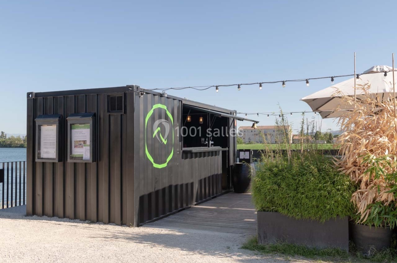 Un conteneur noir aménagé en kiosque avec une terrasse, des plantes et des guirlandes lumineuses, près d'un plan d'eau.