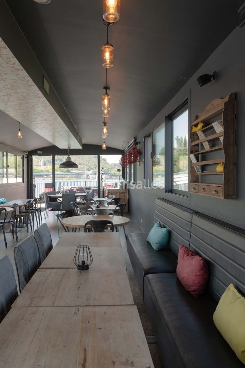 Salle de restaurant lumineuse avec tables en bois, banquettes grises, coussins colorés et suspensions industrielles.