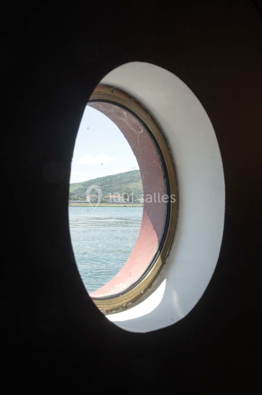 Vue sur une colline verdoyante et un bâtiment au bord de l'eau, encadrée par un hublot circulaire.