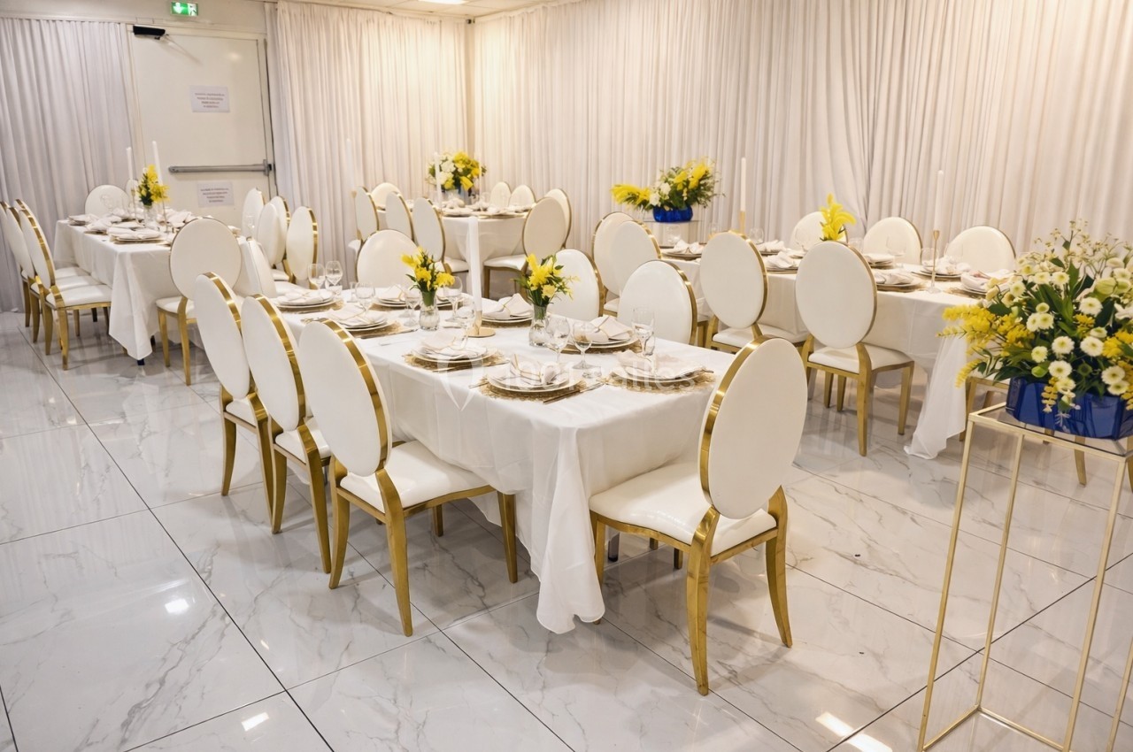 Salle de réception avec tables dressées, nappes blanches, chaises dorées et arrangements floraux jaunes et blancs.