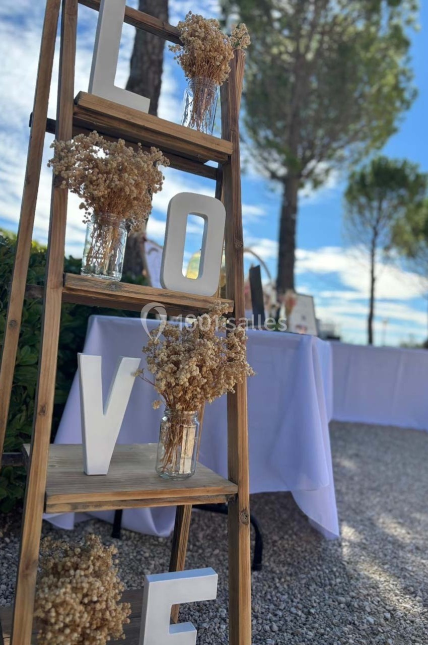 Échelle en bois décorée de lettres formant ’LOVE’ et de bouquets de fleurs séchées, avec un fond extérieur arboré.