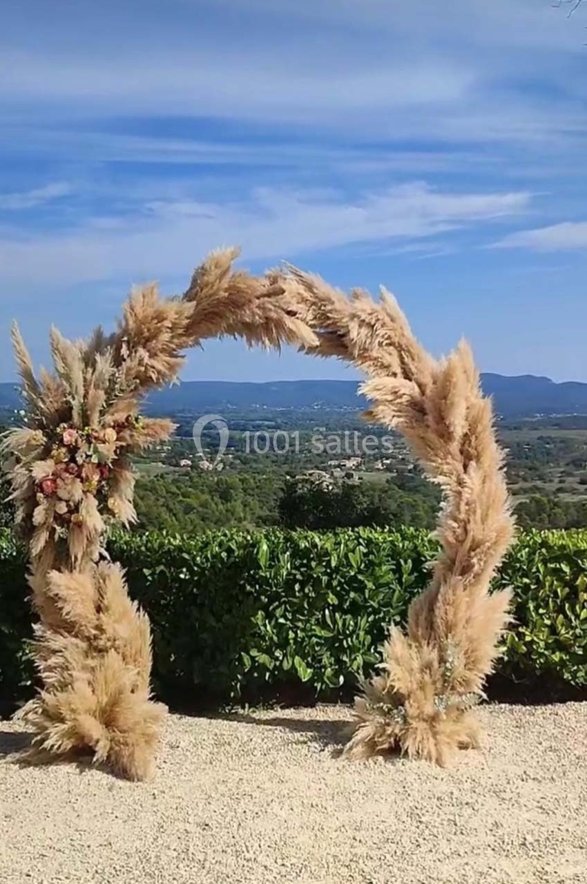 Arche décorative en pampas avec des fleurs, installée en extérieur devant un paysage vallonné sous un ciel bleu.