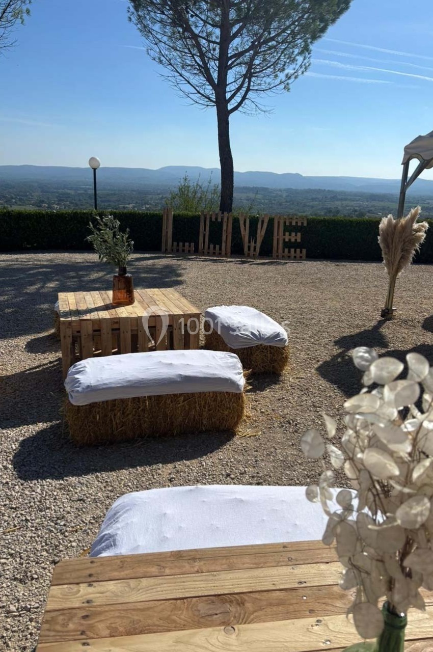 Espace extérieur décoré avec des bottes de paille, palettes en bois, et vue sur un paysage vallonné sous un ciel dégagé.