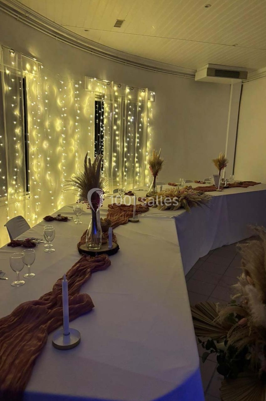 Salle décorée pour un événement, avec guirlandes lumineuses, nappes blanches, bougies et compositions florales.