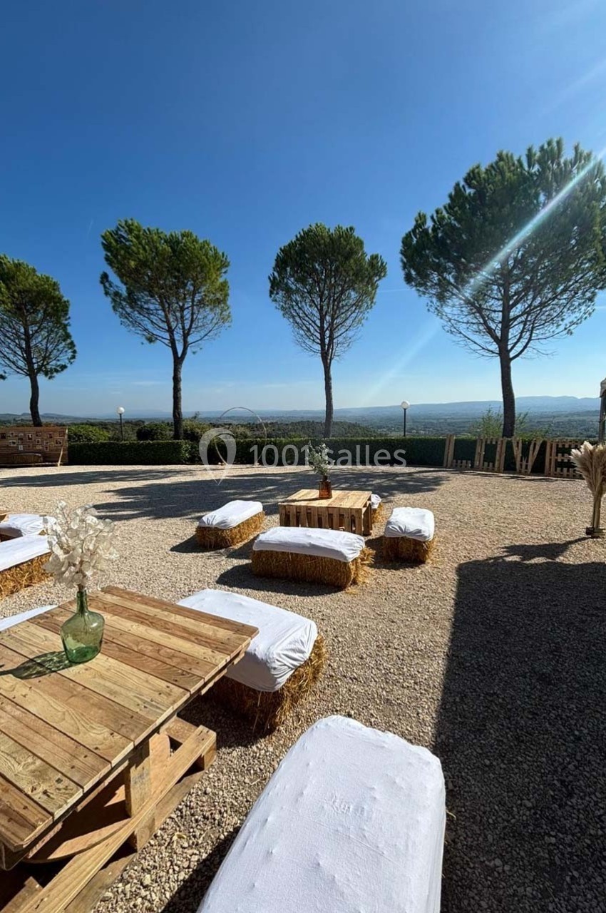 Espace extérieur avec tables en palettes, bottes de paille en sièges, vue dégagée sur un paysage vallonné et arbres en…