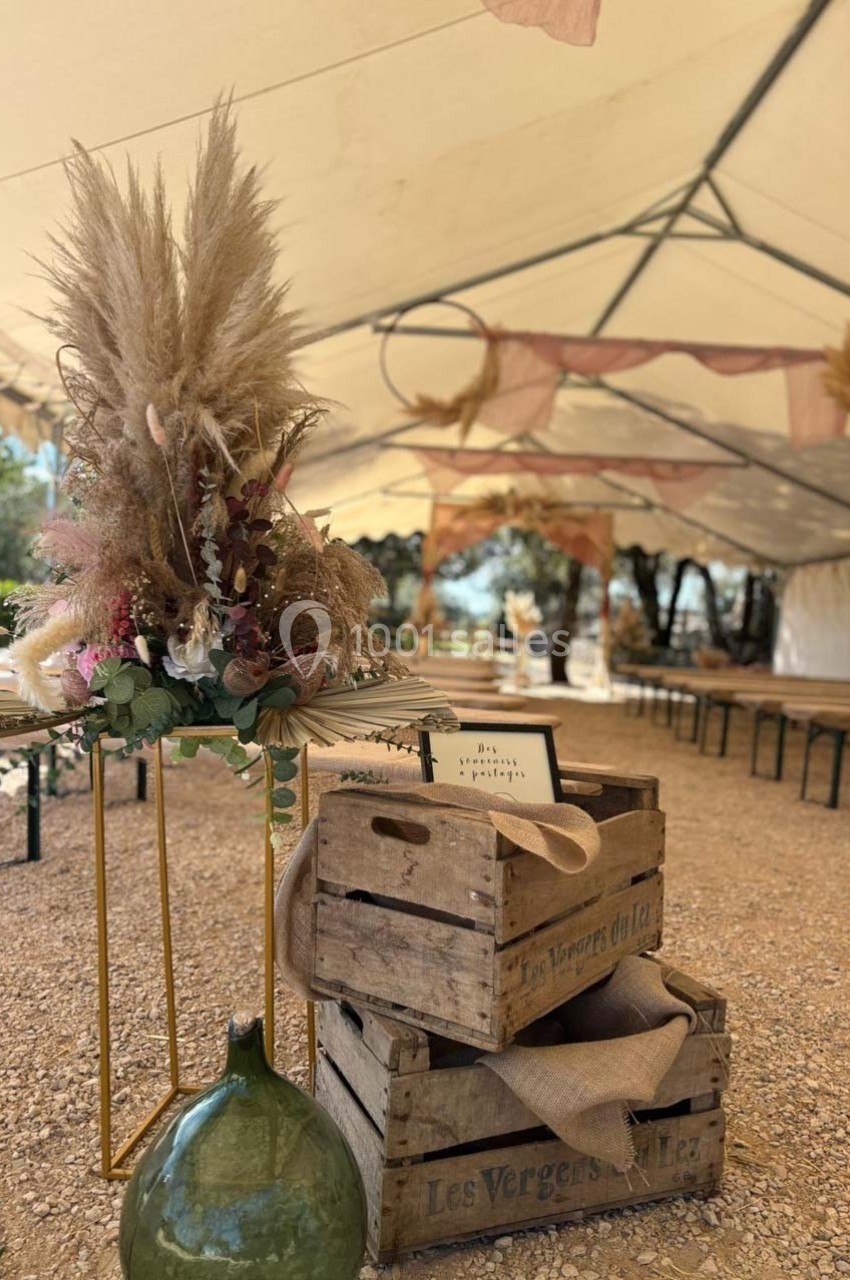 Décoration champêtre avec fleurs séchées, caisses en bois et jarre en verre sous une tente ouverte.