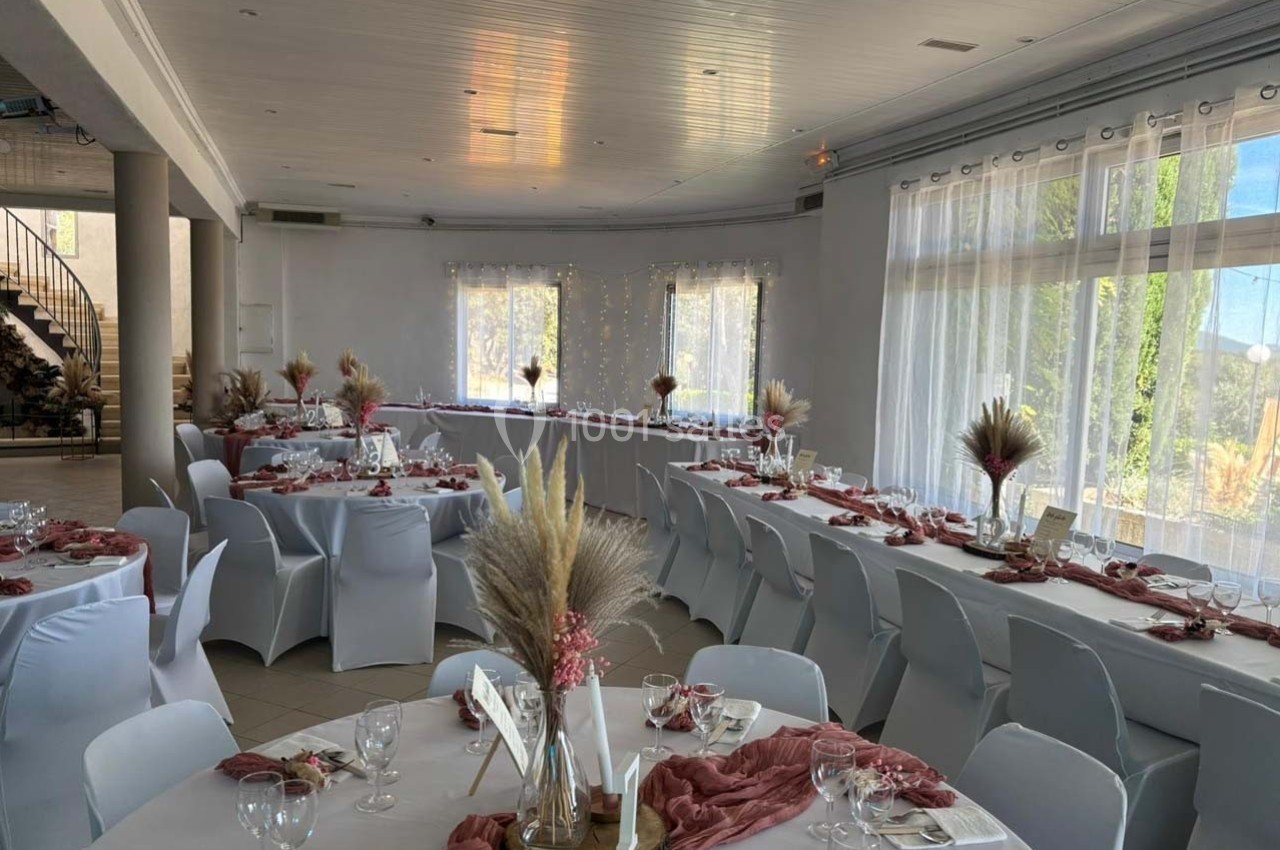 Salle de réception décorée avec des tables rondes et rectangulaires, nappes blanches et ornements floraux dans des tons…