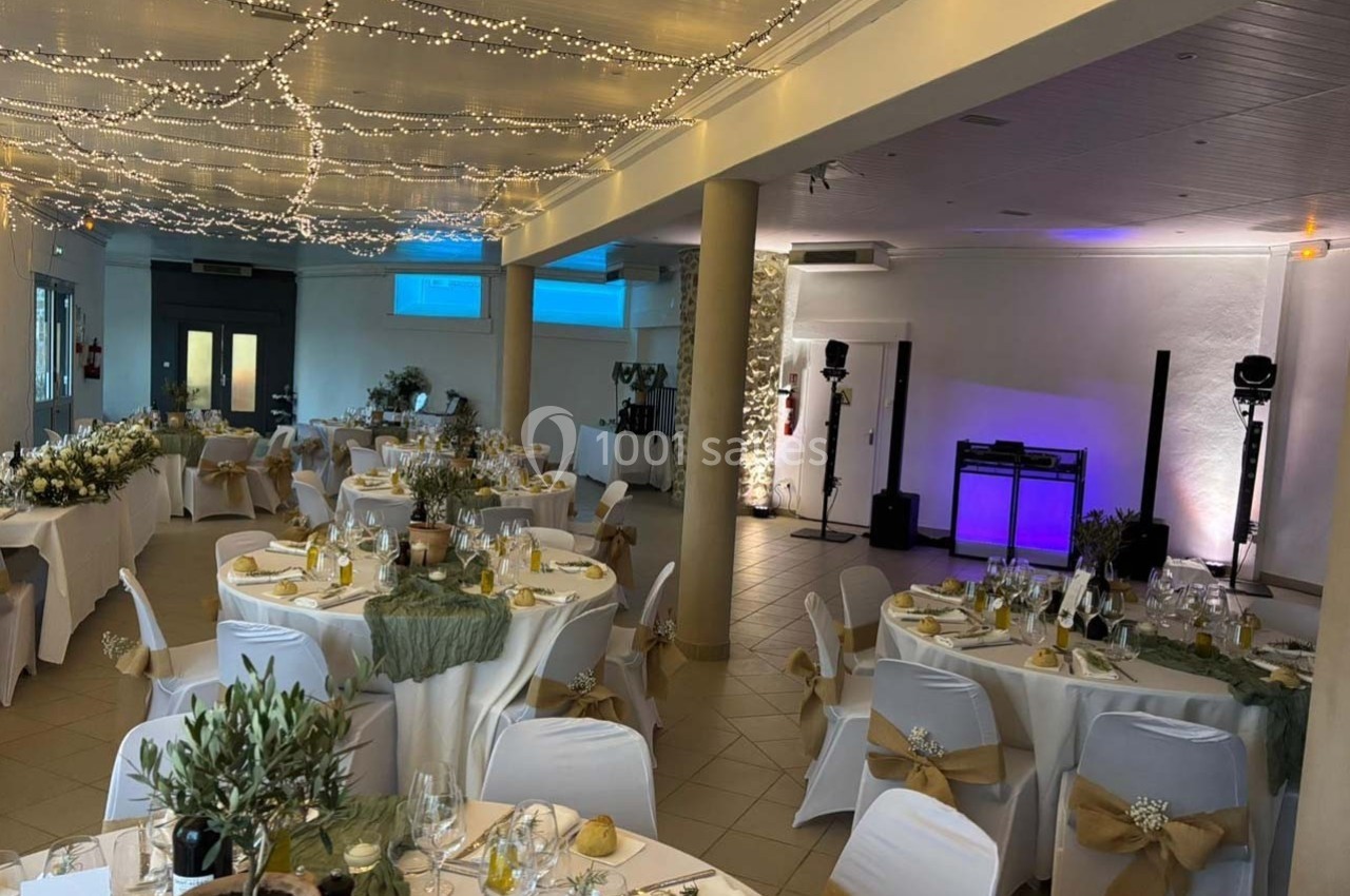 Salle de réception décorée pour un mariage, avec tables dressées, guirlandes lumineuses au plafond et espace DJ éclairé.