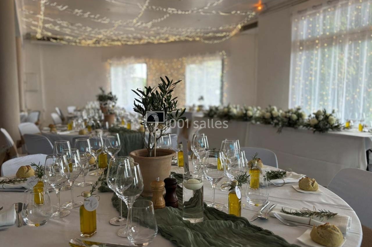 Salle de réception décorée avec des tables dressées, nappes blanches, chemins de table verts et lumières suspendues.