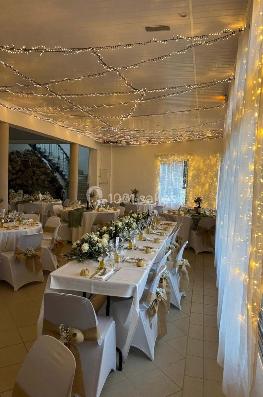 Salle de réception décorée pour un mariage, avec tables dressées, chaises ornées et guirlandes lumineuses au plafond.