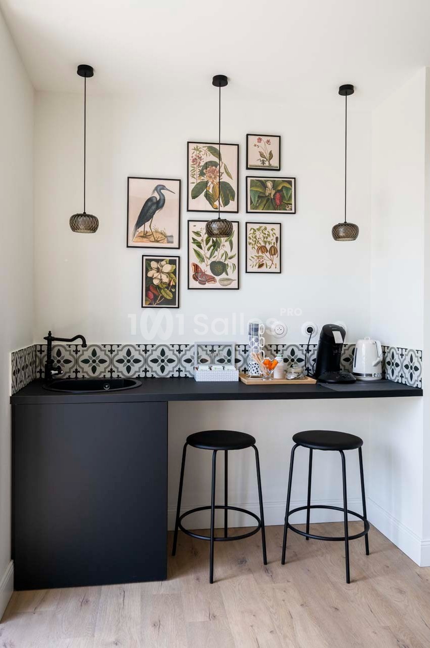Coin kitchenette avec mini-réfrigérateur noir, deux tabourets, cafetière, bouilloire et décorations murales végétales.