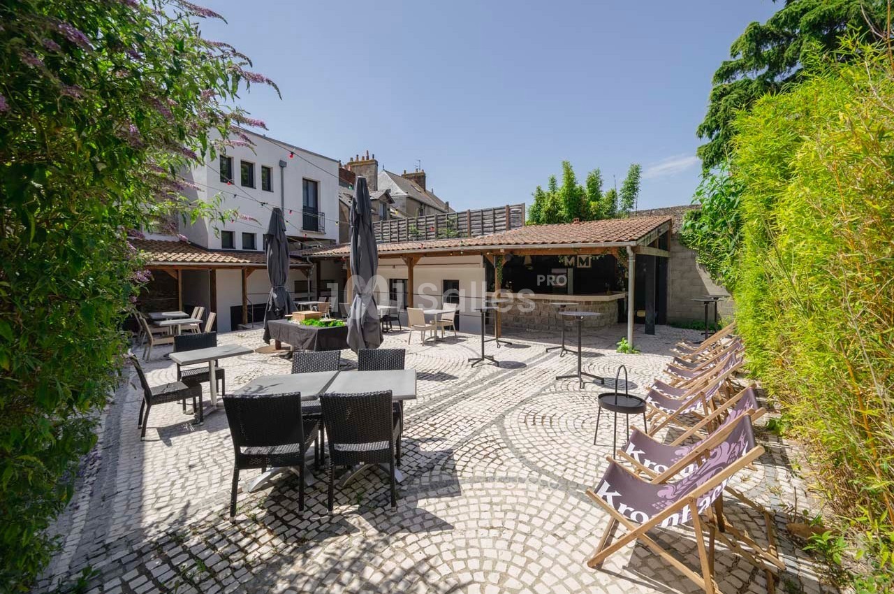 Terrasse extérieure pavée avec tables, chaises, parasols noirs et transats entourée de verdure et bâtiments.