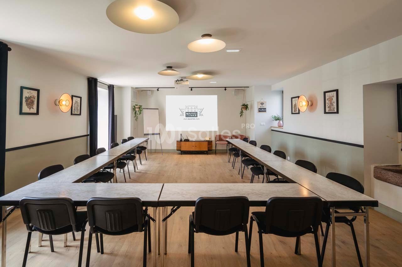 Salle de réunion lumineuse avec disposition en U, tables et chaises noires, écran de projection et décorations murales.