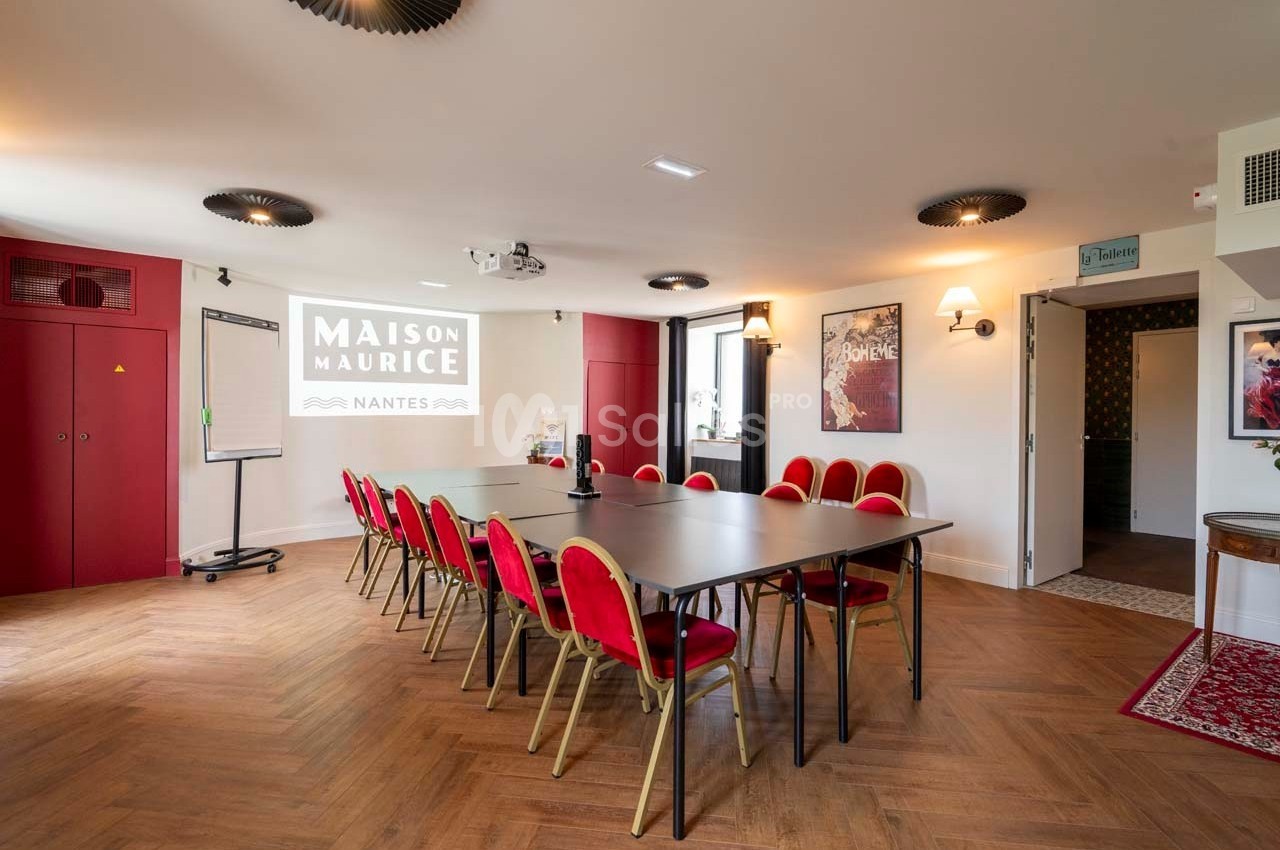 Salle de réunion lumineuse avec table rectangulaire, chaises rouges, projecteur et écran affichant ’Maison Maurice’.