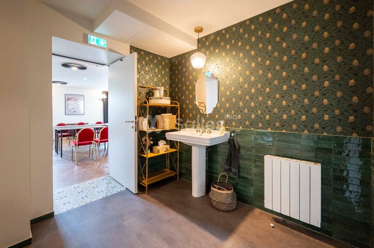 Salle de bain avec lavabo, miroir, étagère en osier, mur vert à motifs et vue sur une salle de réunion adjacente.