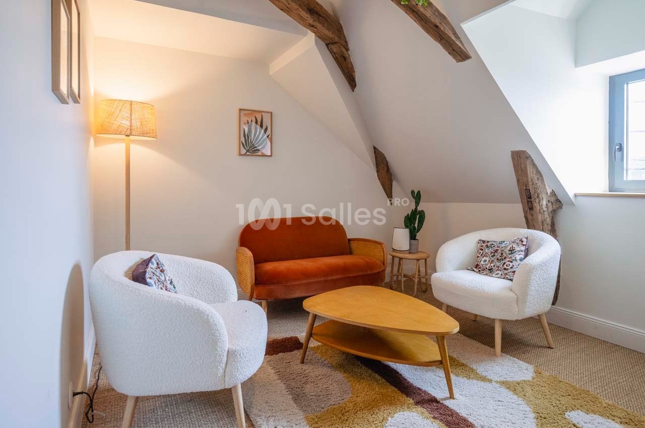 Salon mansardé avec canapé orange, deux fauteuils blancs, table basse en bois, tapis coloré et lampadaire allumé.
