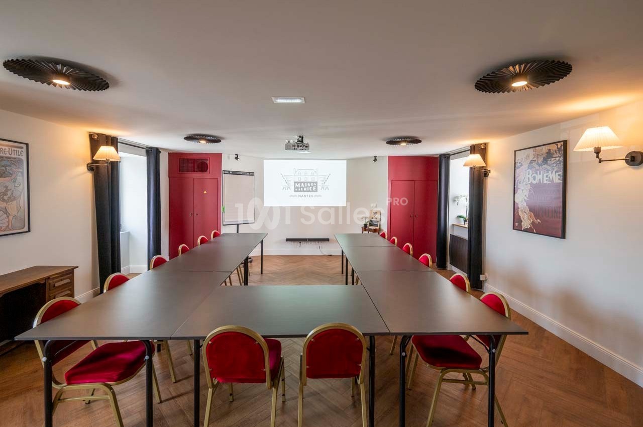 Salle de réunion lumineuse avec tables disposées en U, chaises rouges et écran de projection au fond.